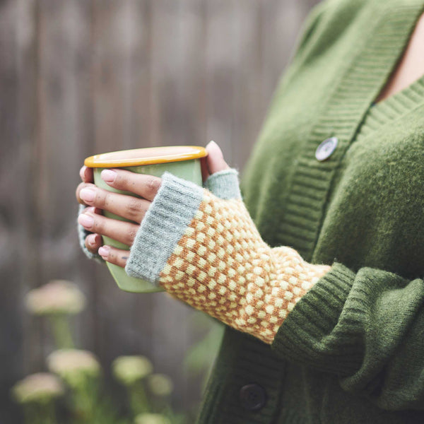 Person wearing a green sweater and patterned fingerless gloves holding a mug outdoors.