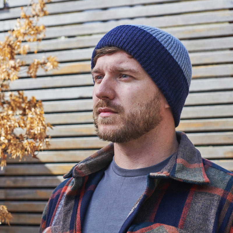 Man wearing a blue knit beanie and plaid jacket standing against a wooden wall.