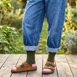 Person wearing blue jeans, green socks, and brown sandals with pink accents outdoors.