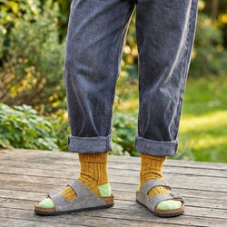 Person wearing mustard yellow socks with grey sandals on a wooden deck with a garden background