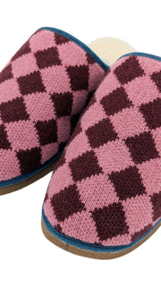A pair of Catherine Tough knit slip-on slippers with a pink and dark burgundy argyle pattern, featuring a cream-colored sheepskin lining and a blue trim along the edge.