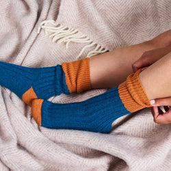 A person wearing a pair of ribbed ankle socks in teal blue and burnt orange, shown sitting on a beige textured blanket.